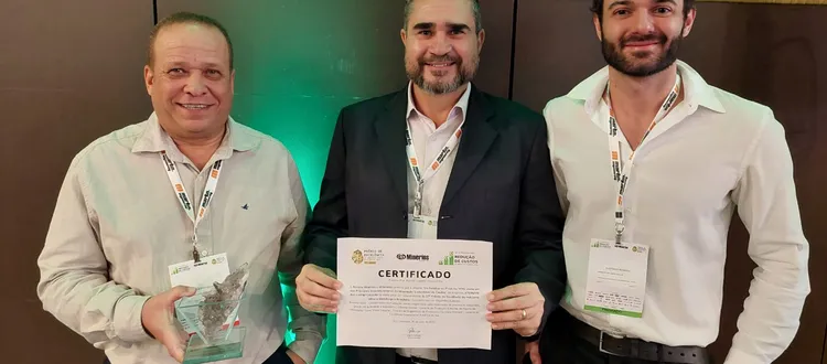 Three men smiling, one holding a certificate and another holding a glass award, wearing lanyards at an indoor event.