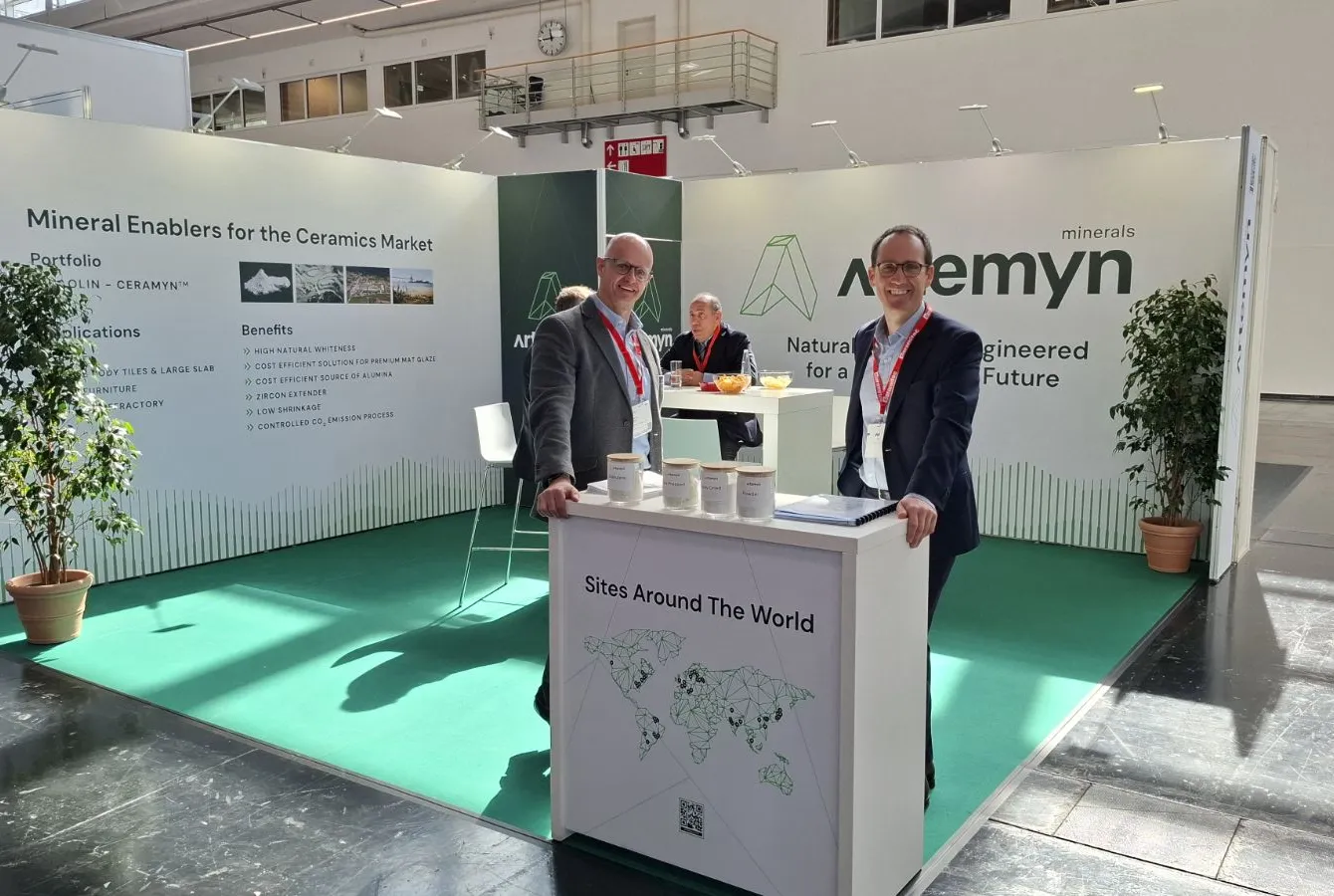Two people stand at an exhibition booth for "Ceramyn," displaying mineral solutions for ceramics. The booth features a world map and benefits list.