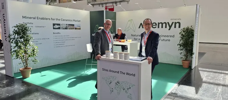 Two people stand at an exhibition booth for "Ceramyn," displaying mineral solutions for ceramics. The booth features a world map and benefits list.