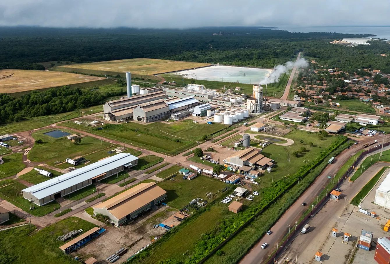 Aerial view of an industrial complex with smoke-emitting chimneys, surrounded by greenery and adjacent to a residential area and roads.