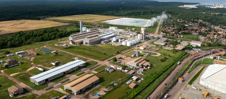 Aerial view of an industrial complex with smoke-emitting chimneys, surrounded by greenery and adjacent to a residential area and roads.