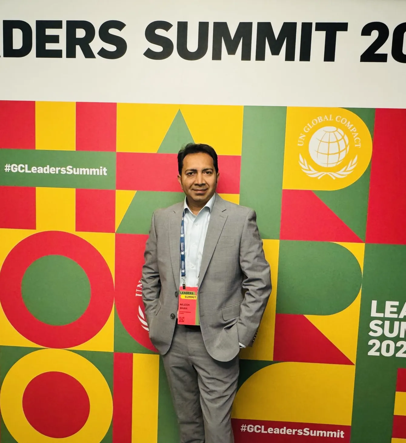 Man in a gray suit stands in front of a colorful "Leaders Summit 2025" backdrop with hashtags and UN Global Compact logos.