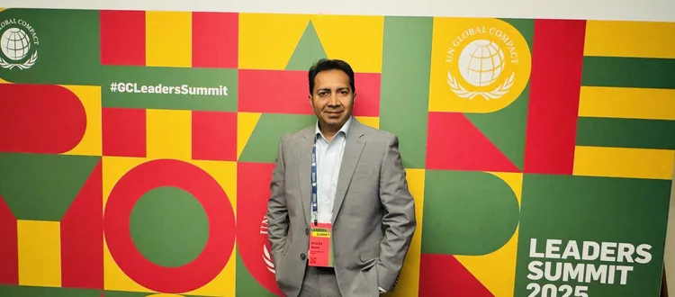 Man in a gray suit stands in front of a colorful "Leaders Summit 2025" backdrop with hashtags and UN Global Compact logos.