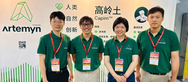 Four people in green Artemyn shirts with red lanyards pose at a trade booth featuring the company's logo and product information.