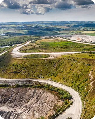 Aerial view of a winding road through green hills and a quarry, with expansive landscapes stretching to the horizon under a cloudy sky.