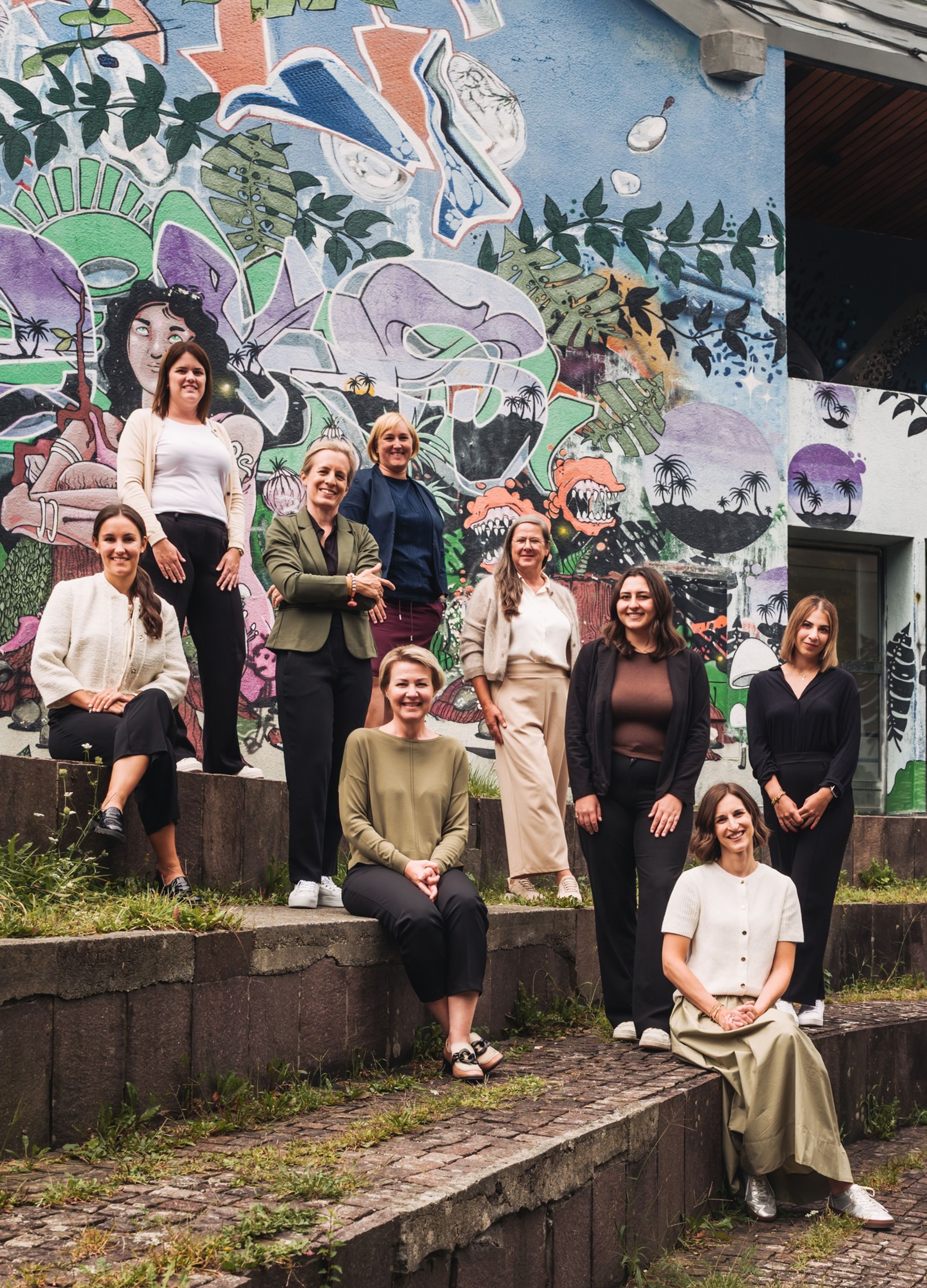 Neun Frauen posieren auf Stufen vor einem farbenfrohen Wandgemälde mit Pflanzen und Tieren, gekleidet in Freizeit- und Geschäftskleidung, und lächeln herzlich.