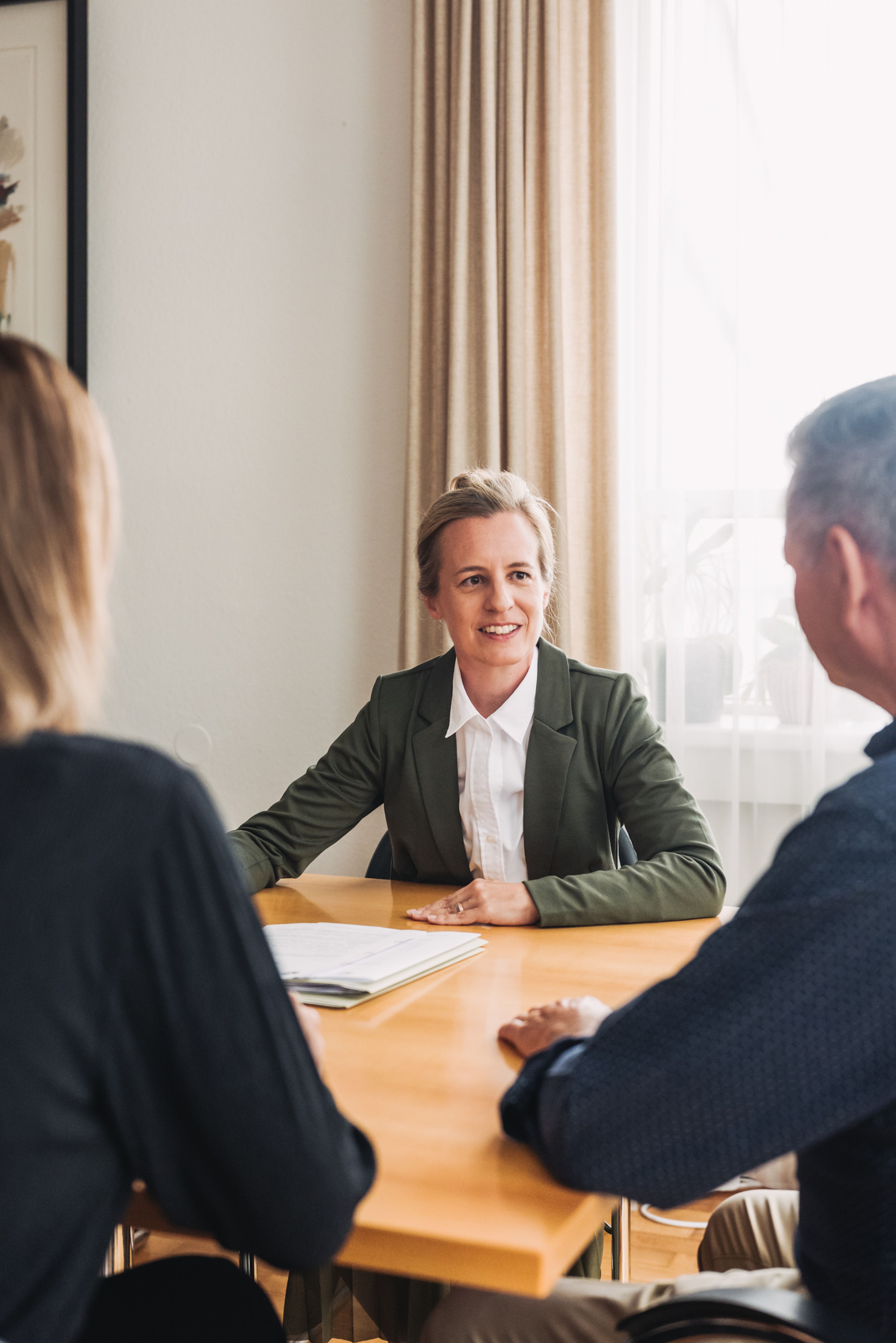 Eine Frau in einem grünen Blazer sitzt an einem Tisch, lächelt und unterhält sich mit zwei Personen gegenüber von ihr in einer Büroumgebung mit sanftem Tageslicht.