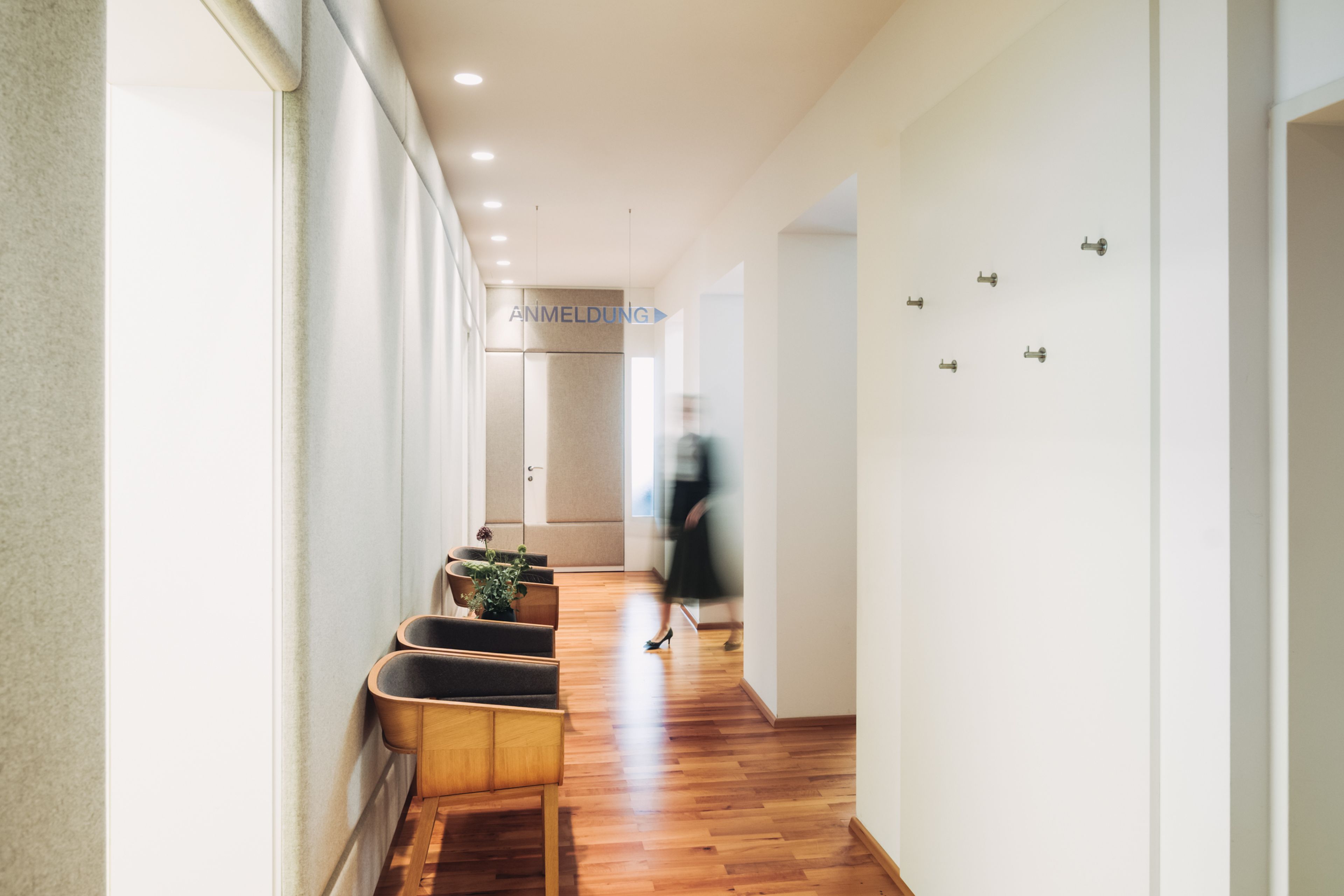 Ein moderner, gut beleuchteter Flur mit Holzboden, gepolsterten Stühlen und einer Person, die auf eine Tür unter einem Schild mit der Aufschrift „ANMELDUNG” zugeht.