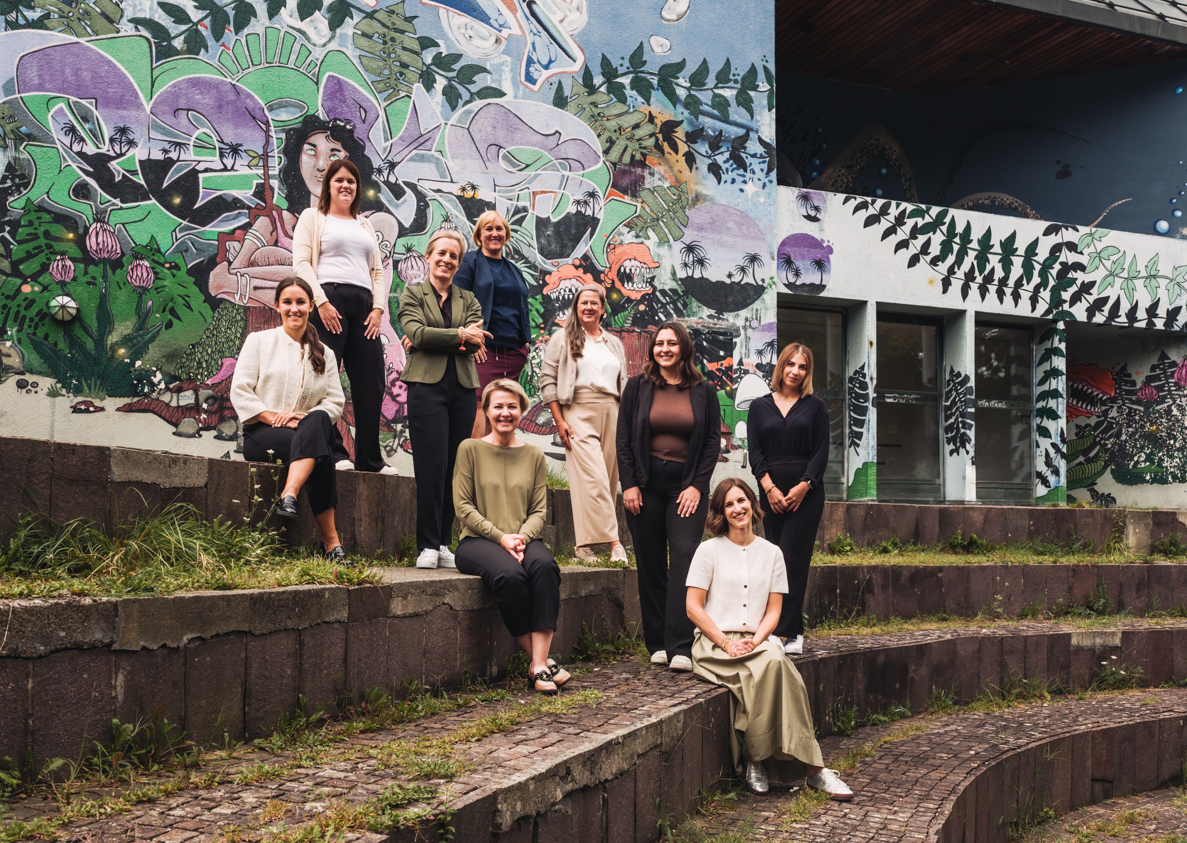 Neun Personen sitzen und stehen auf Stufen vor einer bunten, mit Graffiti bedeckten Wand.