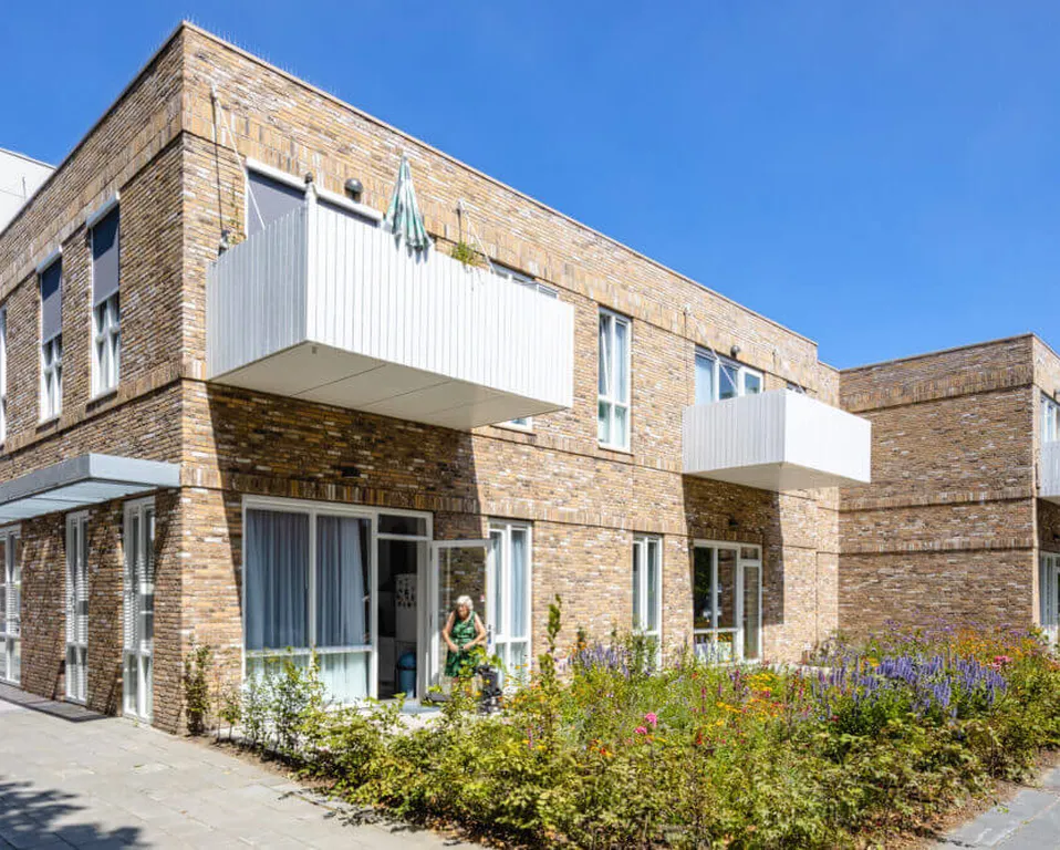 Modern bakstenen appartementencomplex met grote ramen, balkons en een kleine tuin. Een persoon loopt op het trottoir onder een helderblauwe lucht.