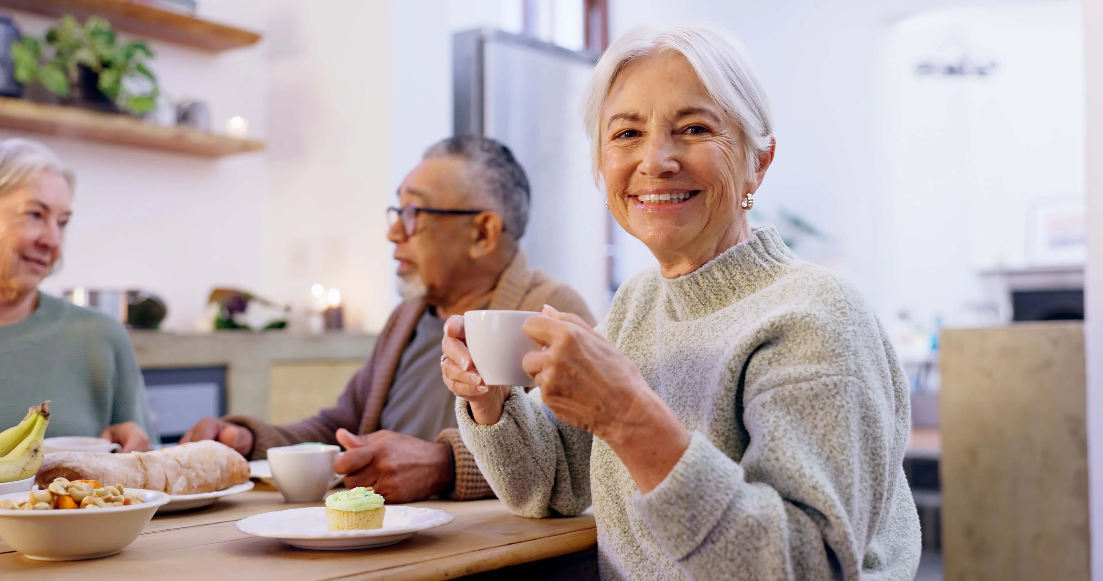 Oudere vrouw glimlacht terwijl ze met een kopje koffie aan tafel zit met andere senioren en geniet van het eten en het gesprek in een gezellige keuken.