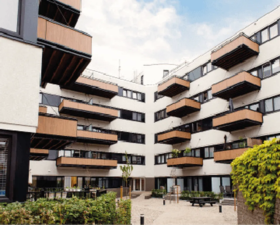 Modern apartment building with wooden balconies, white facade, and a courtyard surrounded by greenery under a partly cloudy sky.