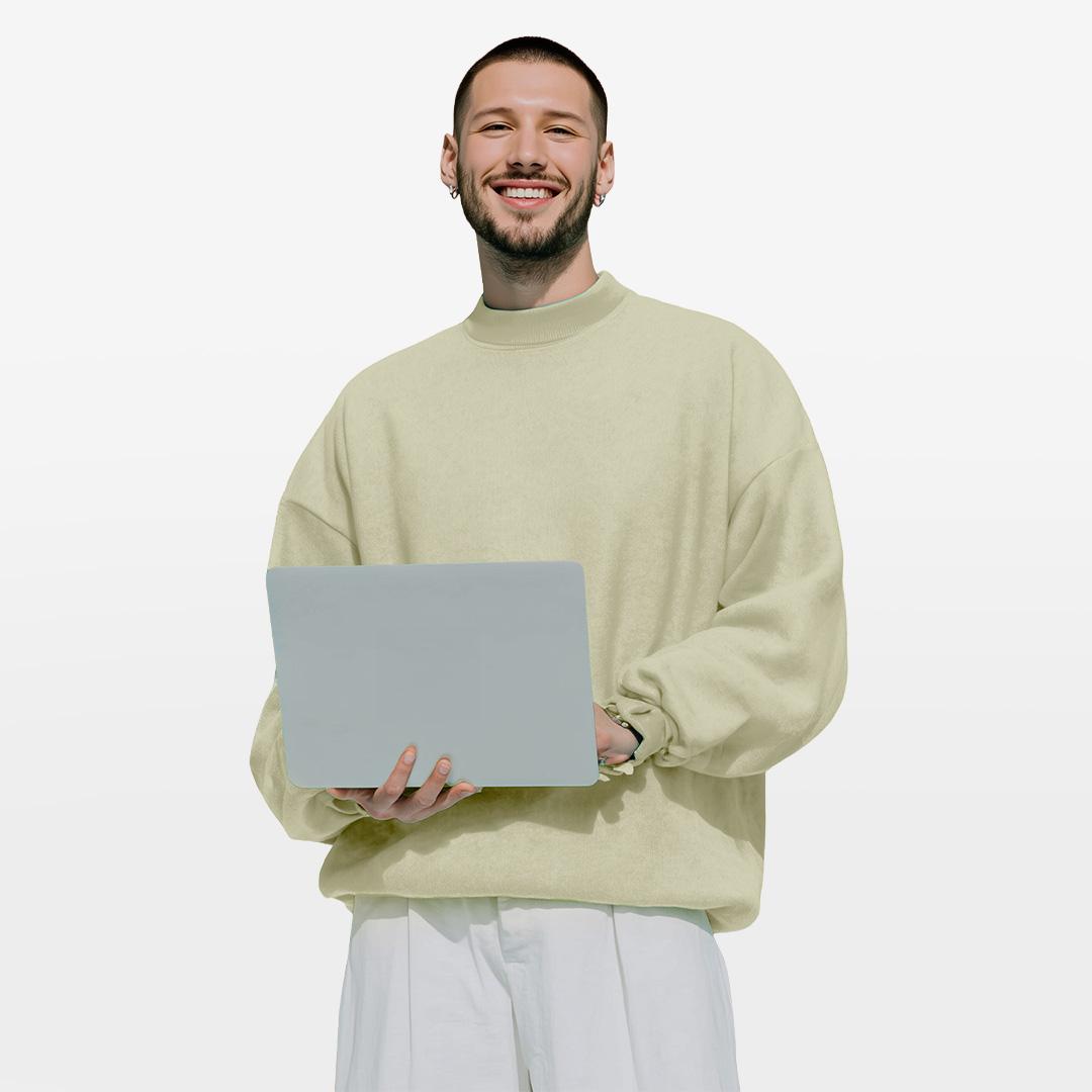 Smiling man in a beige sweater holds a laptop, standing against a plain white background.