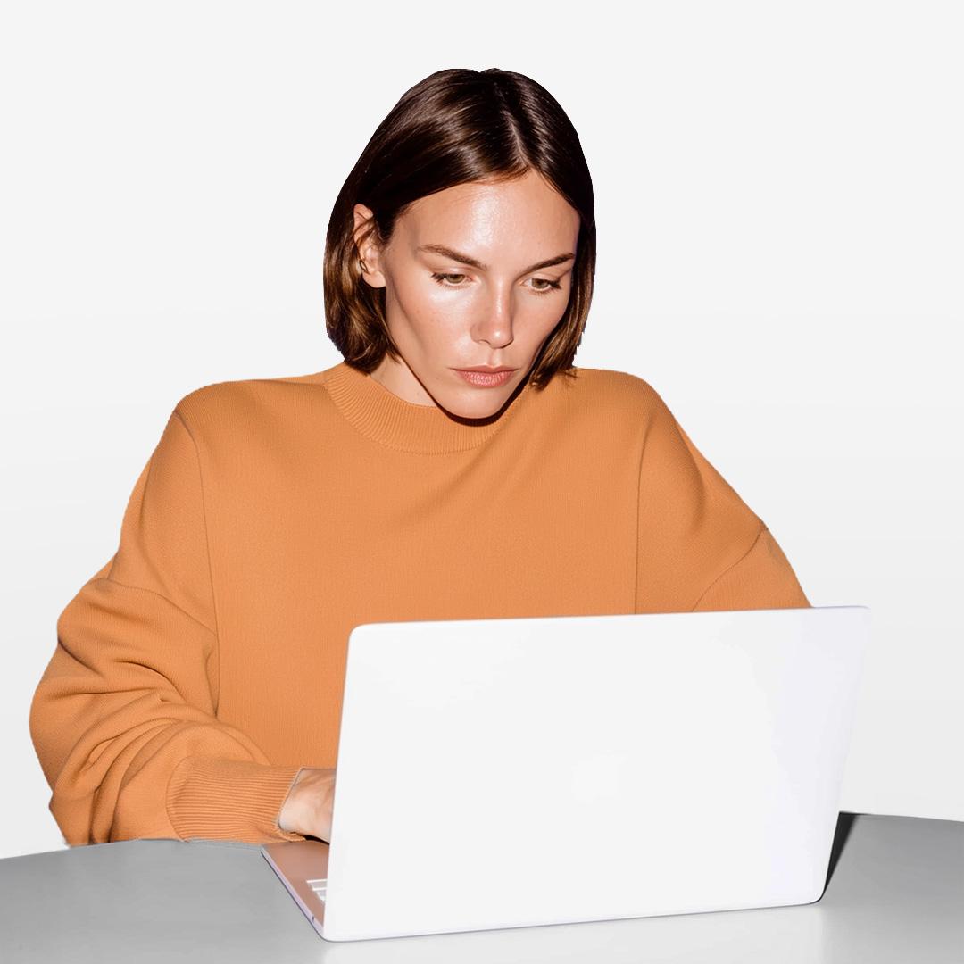 Person with short brown hair in an orange sweater using a white laptop, focused on the screen, against a plain white background.