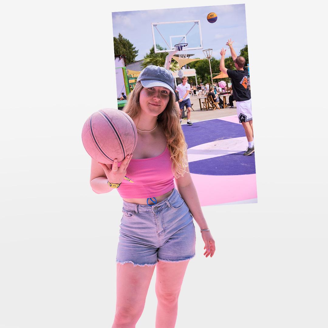 Woman in pink top and denim shorts holding a basketball, standing in front of an outdoor basketball court with players.
