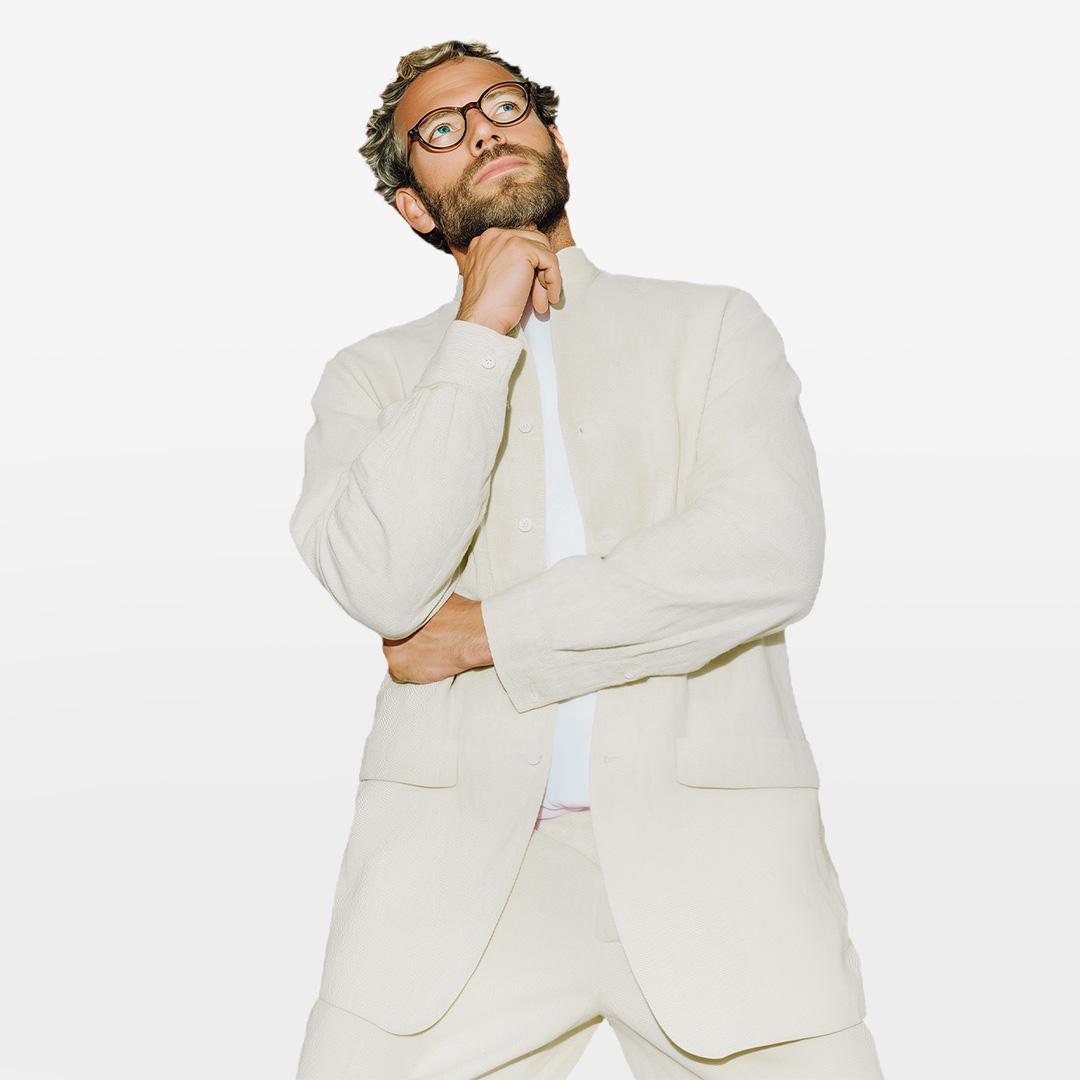 Man with glasses in a white suit poses confidently with a hand on his chin against a plain background.