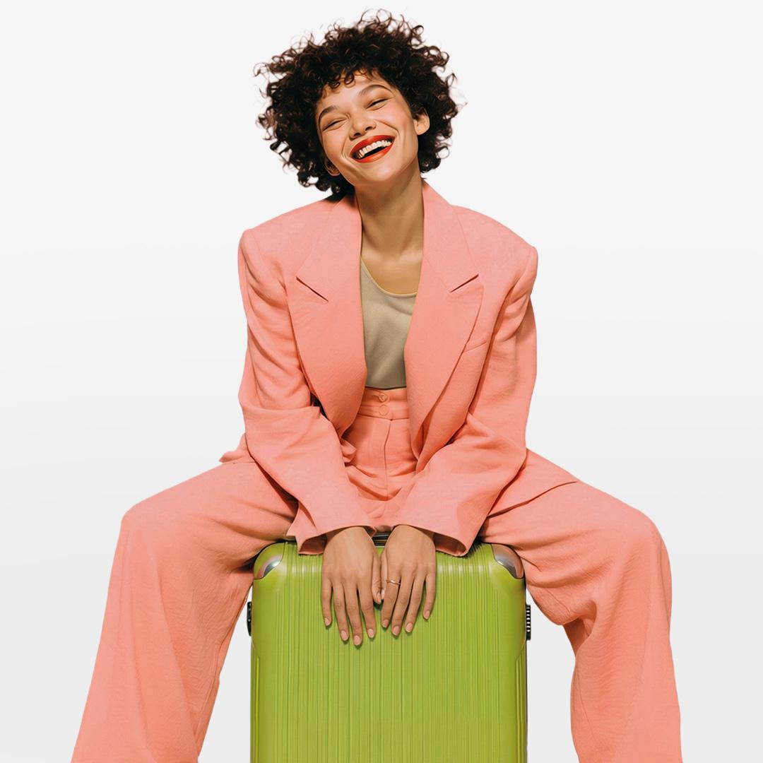 Woman in a pink suit sitting on a green suitcase, smiling with eyes closed against a plain background.