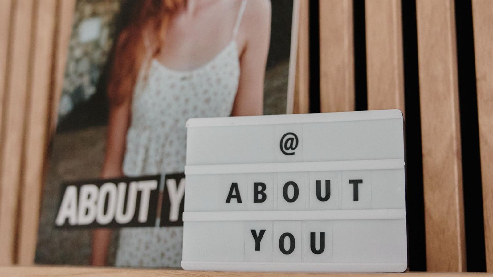 Sign reading "@ About You" on a wooden shelf, with a photo of a red-haired woman in a white dress in the background.