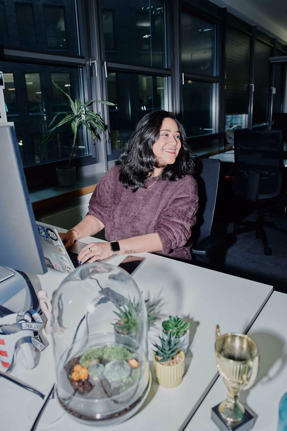 Smiling woman working at a desk with plants and a trophy, inside a modern office at night.