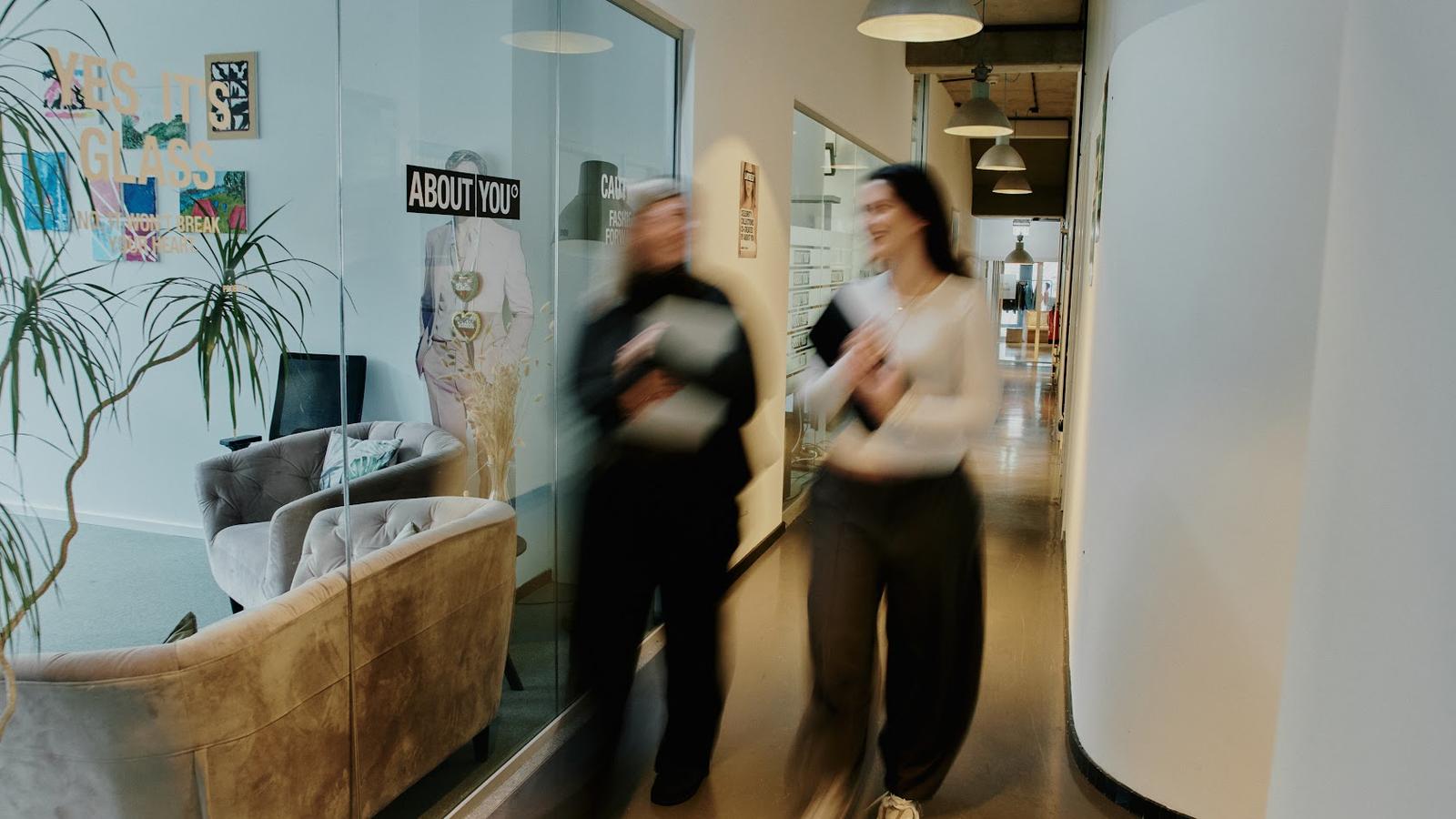 Two people walking briskly down a hallway with glass walls, holding papers. A seating area with plants is visible through the glass.