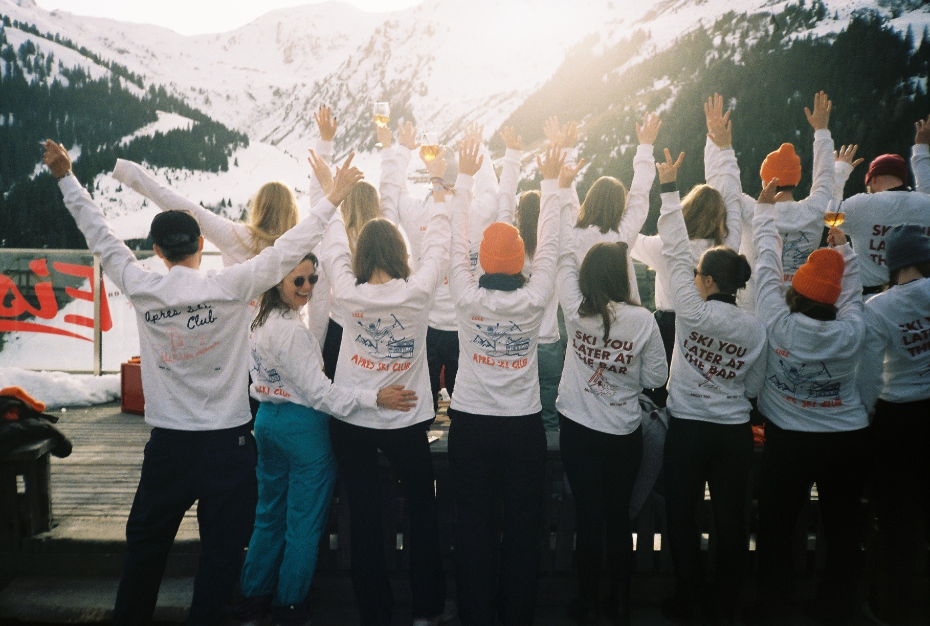 A group of people in matching white jackets and orange hats raise their arms while standing on a snowy mountain deck.