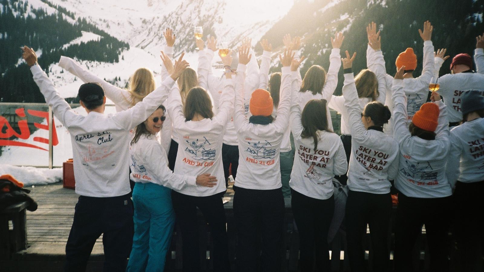 A group of people in matching white jackets and orange hats raise their arms while standing on a snowy mountain deck.