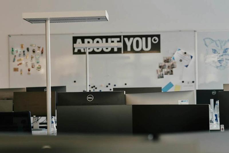 Open-plan office with multiple Dell monitors, whiteboards showing a cat drawing, magnets, and photos. A sign reads "About You" in the background.