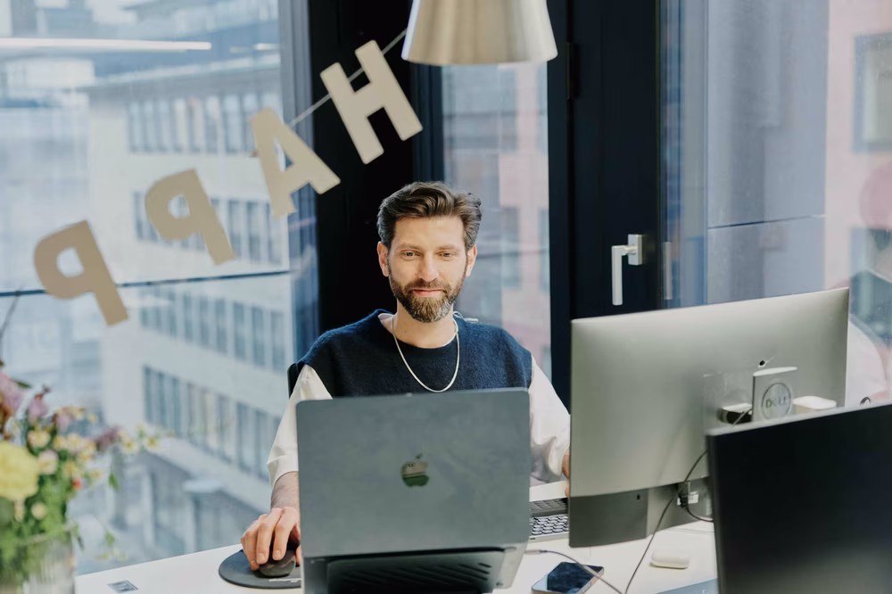 Man with beard working at a desk with dual monitors, in an office with a "HAPPY" banner on the window and flowers nearby.