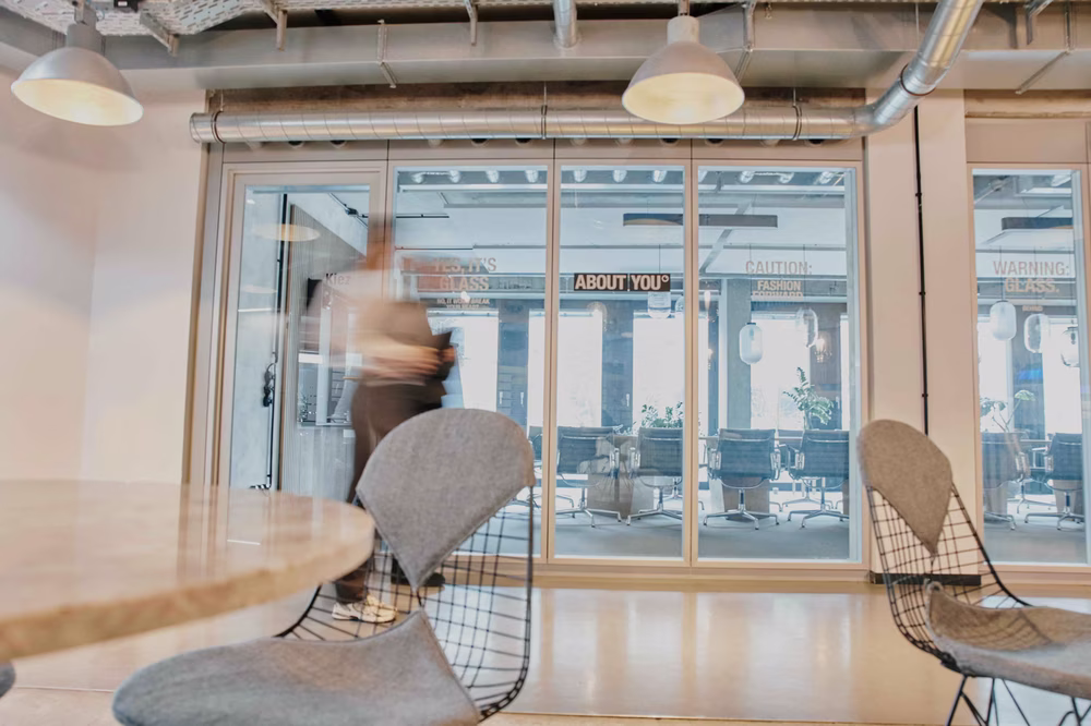 Modern office space with glass wall, blurred person walking, stylish chairs, and overhead lights, creating a dynamic and contemporary atmosphere.