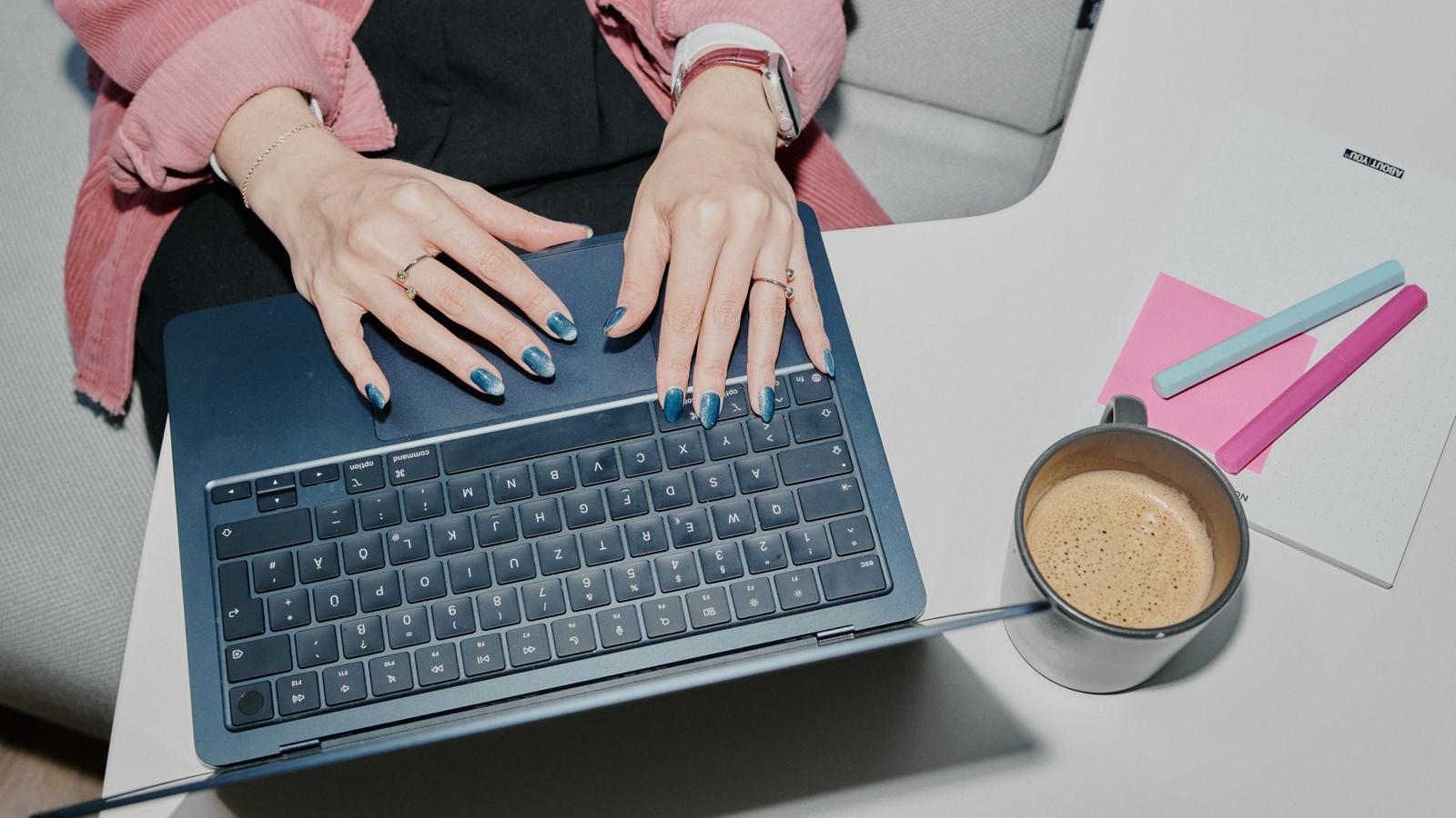 Person typing on a laptop with blue nail polish, next to a coffee cup and notepad with pink and blue pens on a desk.