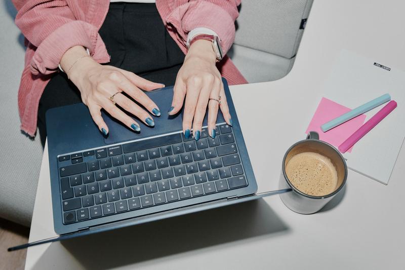 Person typing on a laptop with blue nail polish, next to a coffee cup and notepad with pink and blue pens on a desk.