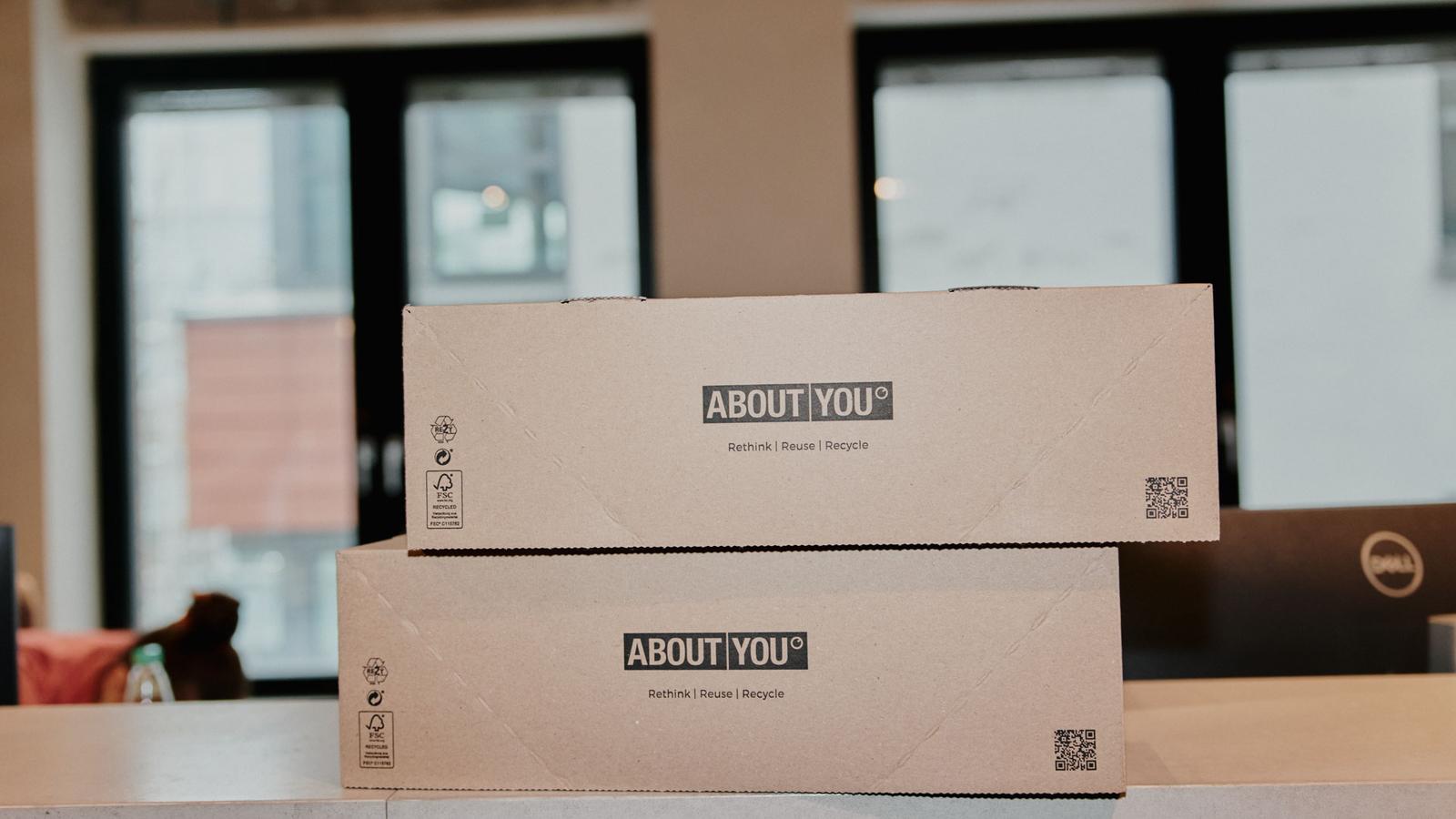 Two stacked cardboard boxes with "ABOUT YOU" branding, placed on a counter in a modern office setting near large windows.