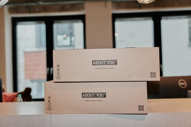 Two stacked cardboard boxes with "ABOUT YOU" branding, placed on a counter in a modern office setting near large windows.