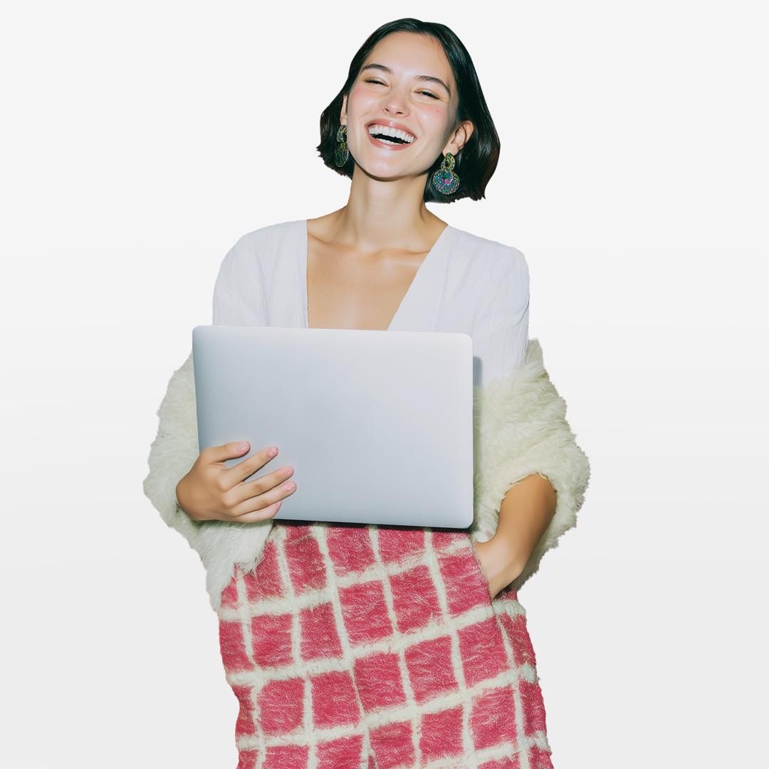 Smiling woman holding a laptop, wearing a white top, fluffy shawl, and red plaid skirt, against a plain background.
