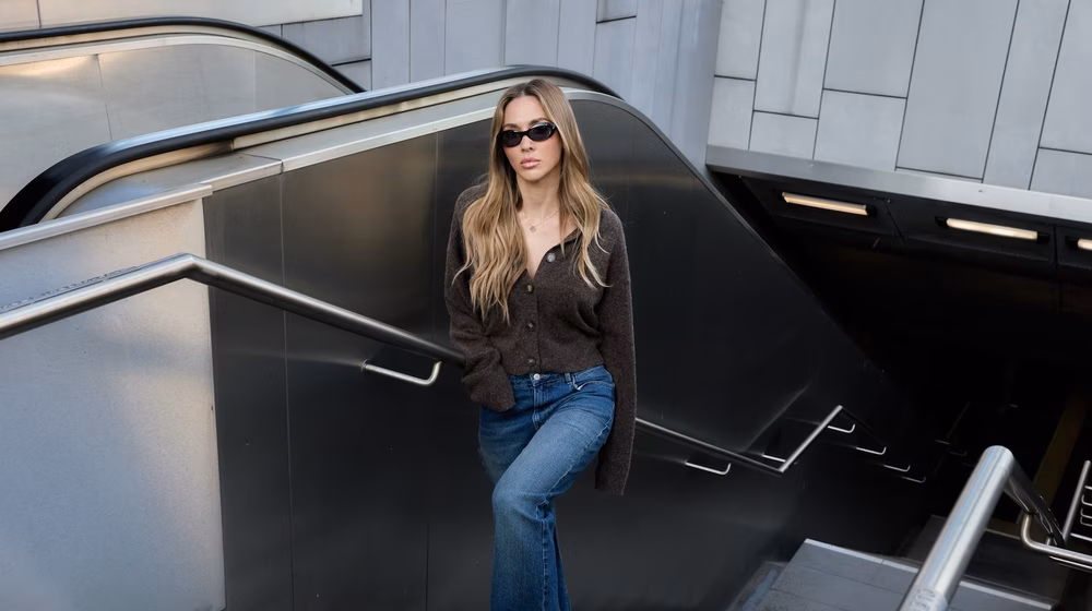 ABOUT YOU a woman with long hair and sunglasses leans against an escalator railing, wearing a dark shirt and jeans, in a modern urban setting.