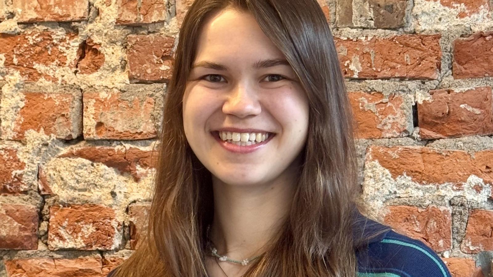 A woman with long brown hair smiles in front of a rustic red brick wall, wearing a striped navy and green shirt.