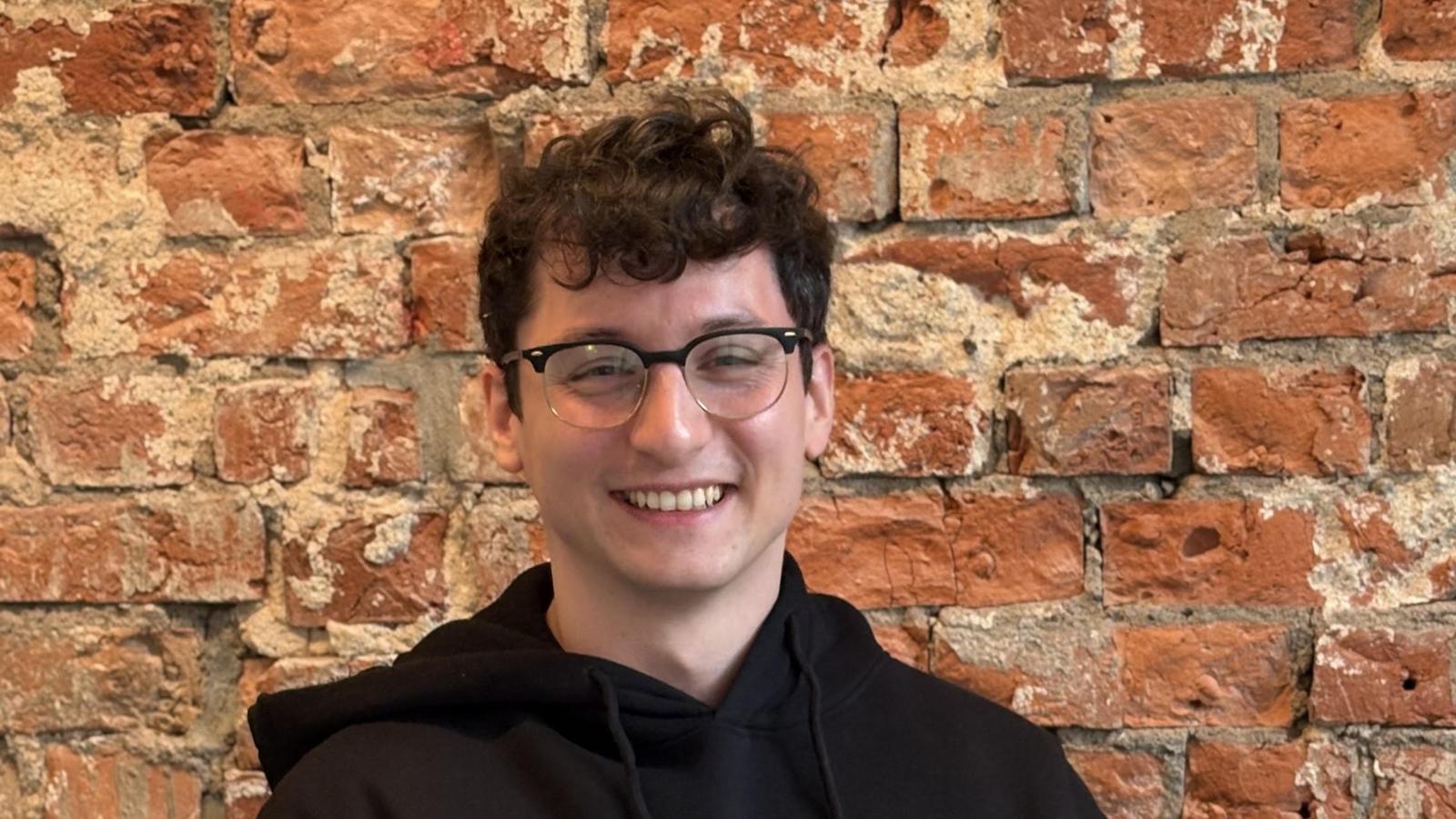 A smiling person wearing glasses and a black hoodie with "About You Company Hoodie" text stands in front of a brick wall.