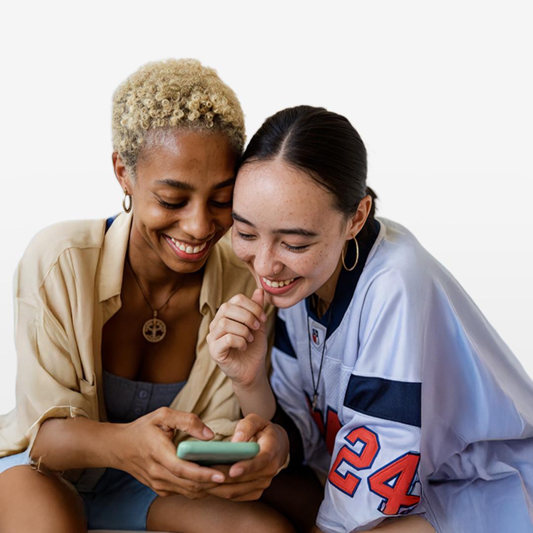 Two people smiling and looking at a smartphone together, sitting closely. One wears a beige shirt, the other a sports jersey.