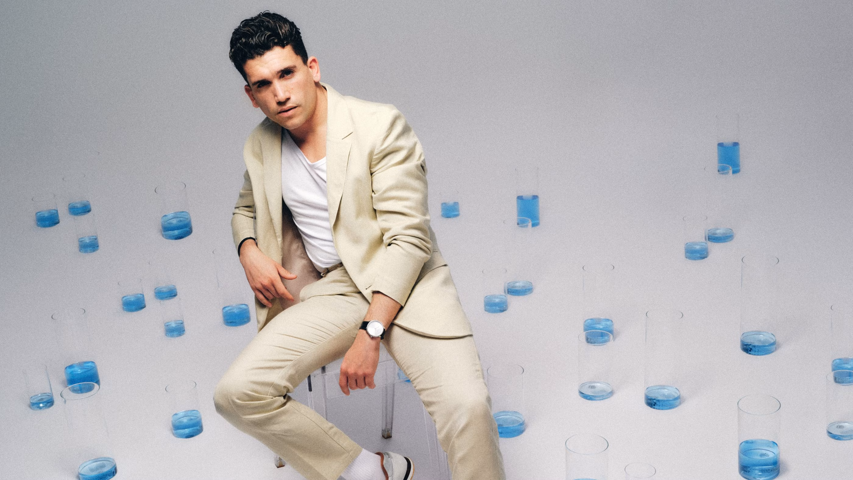 Man in a beige suit sits amid scattered clear glasses filled with blue liquid on a white background.