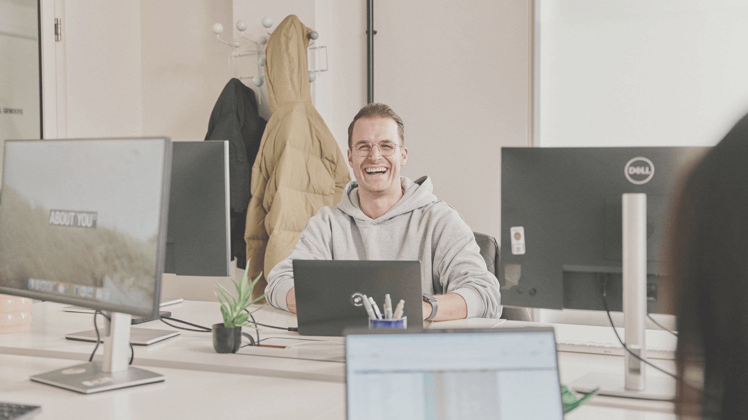 A person in a gray hoodie sits at a desk with multiple monitors, smiling. A coat hangs in the background, and a small plant is on the desk.
