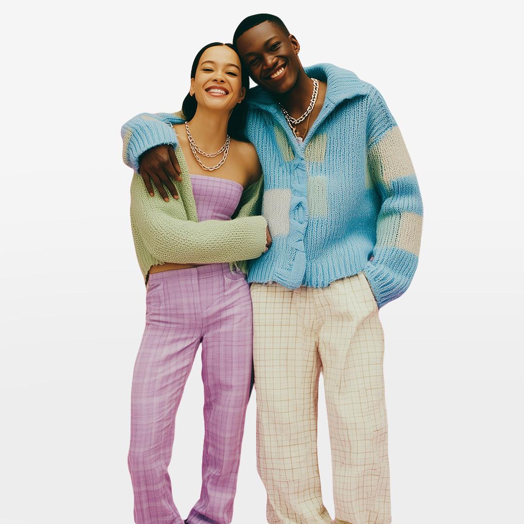 Two people smiling and embracing, wearing colorful knit sweaters and plaid pants, against a plain white background.