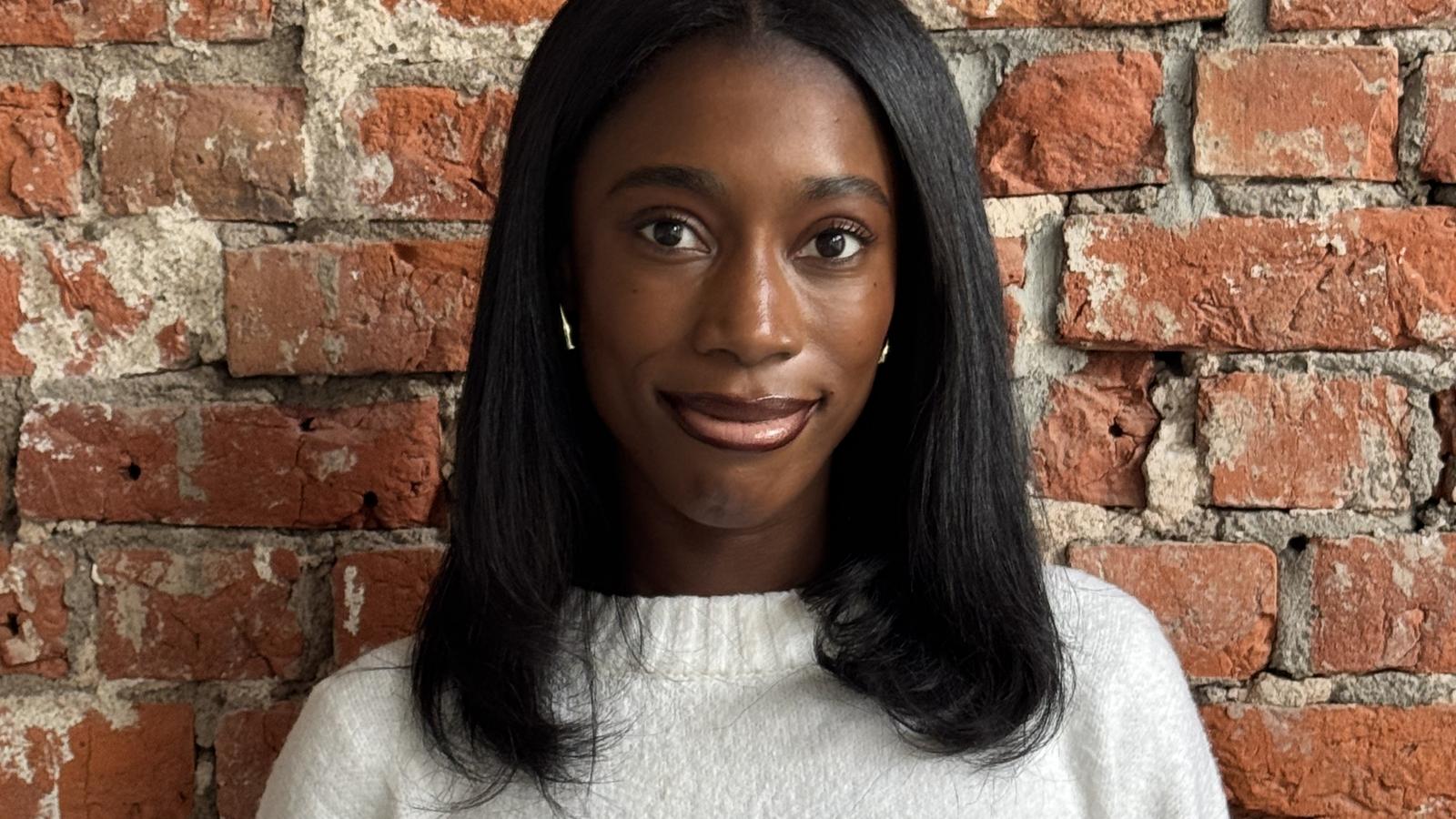 A person in a white sweater stands smiling in front of a rustic red brick wall.