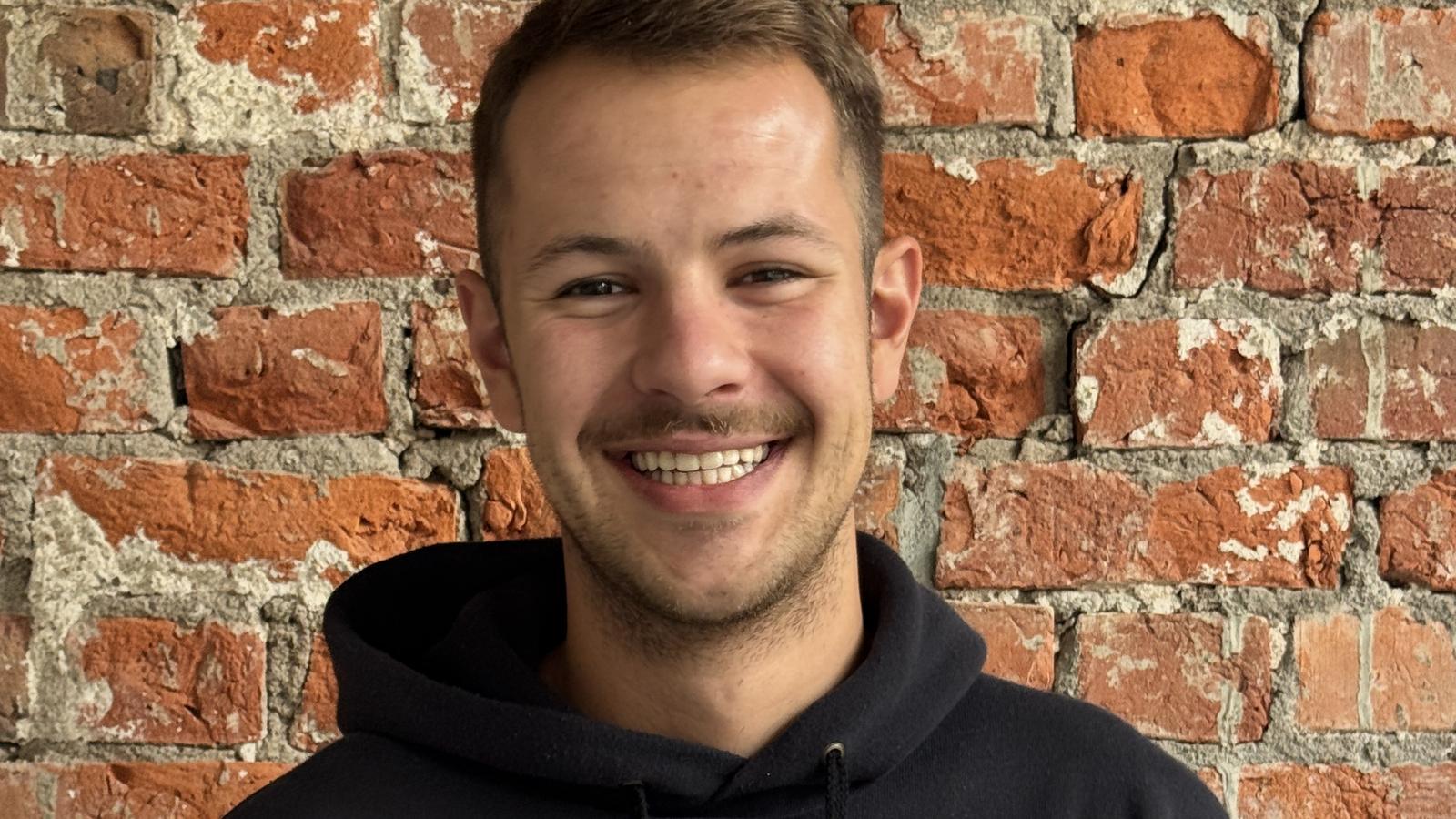 Smiling person wearing a black Ramones hoodie, standing in front of a textured red brick wall.