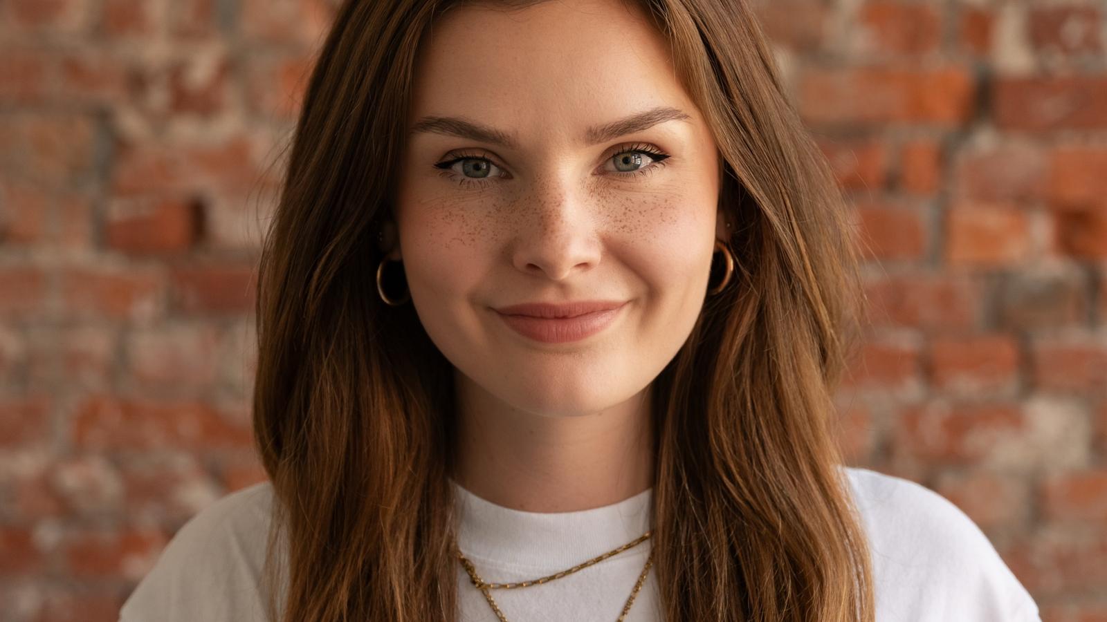 A woman with long brown hair smiles against a brick wall, wearing a white shirt and layered gold necklaces.