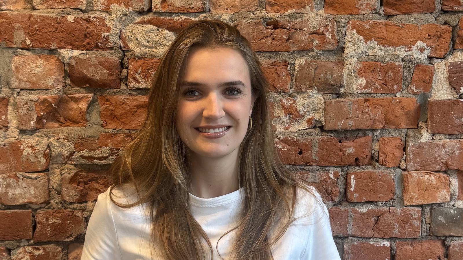 A woman with long hair wearing a white t-shirt stands smiling in front of a red brick wall.
