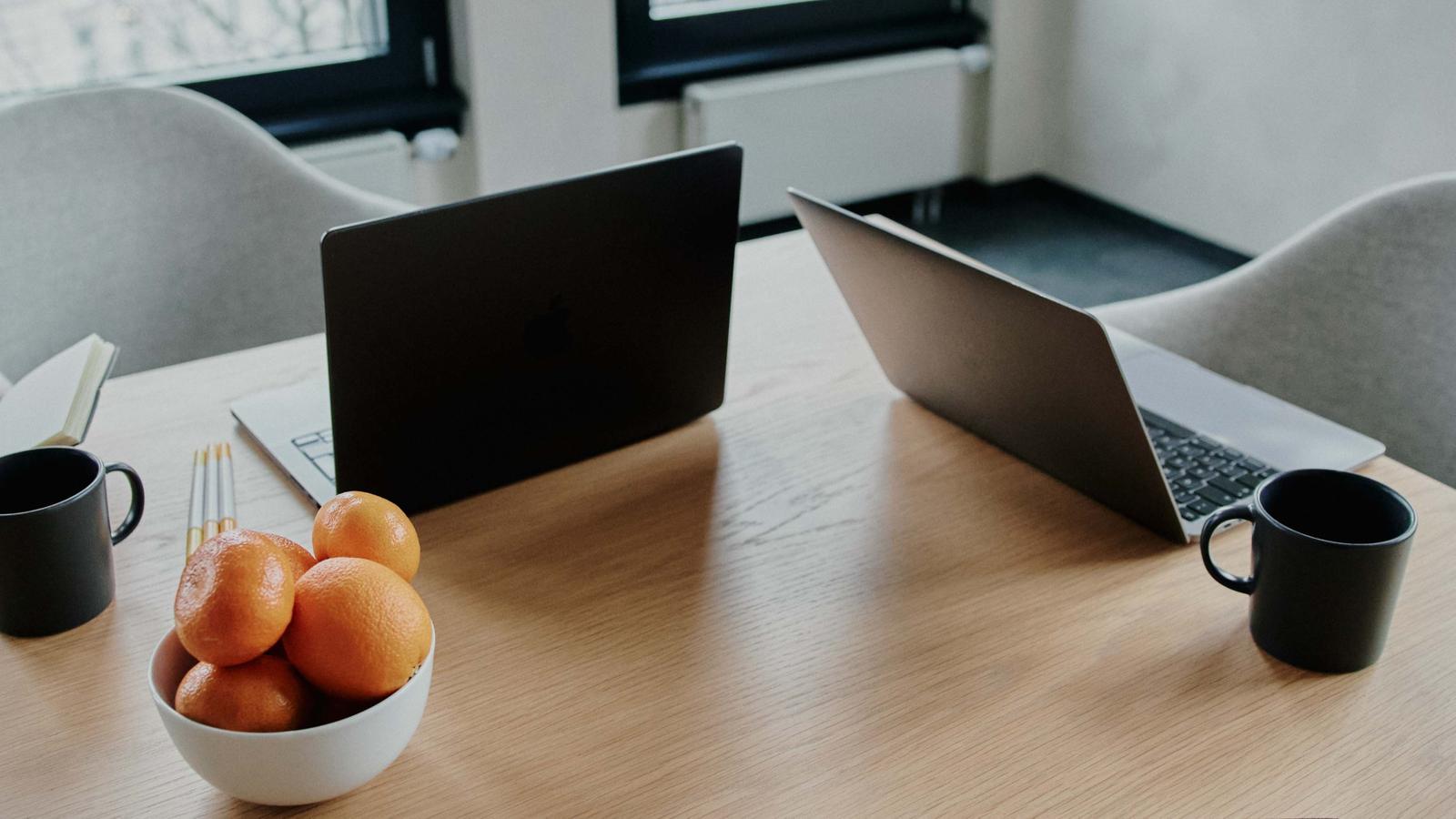 Modern office with two laptops, a bowl of oranges, two black mugs, and a smartphone with a smiley face case on a wooden table.