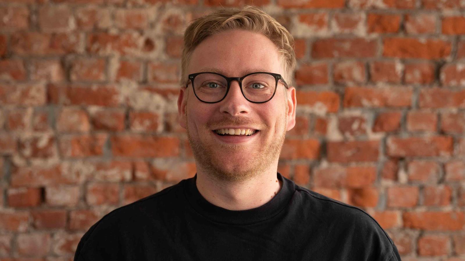 A smiling person with glasses and a black shirt stands against a red brick wall.