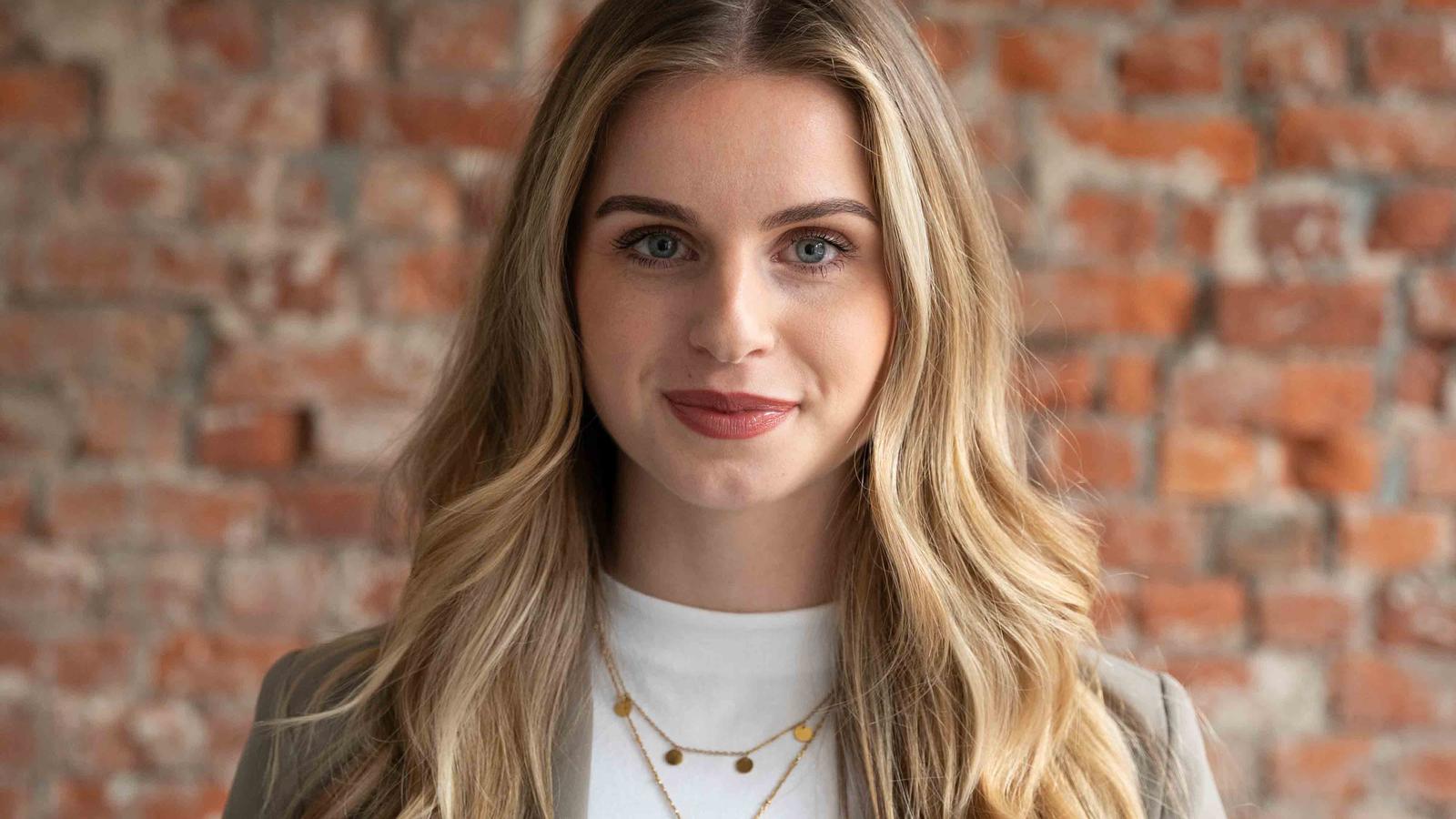 Woman with long blonde hair, wearing a gray blazer and necklaces, stands against a brick wall.
