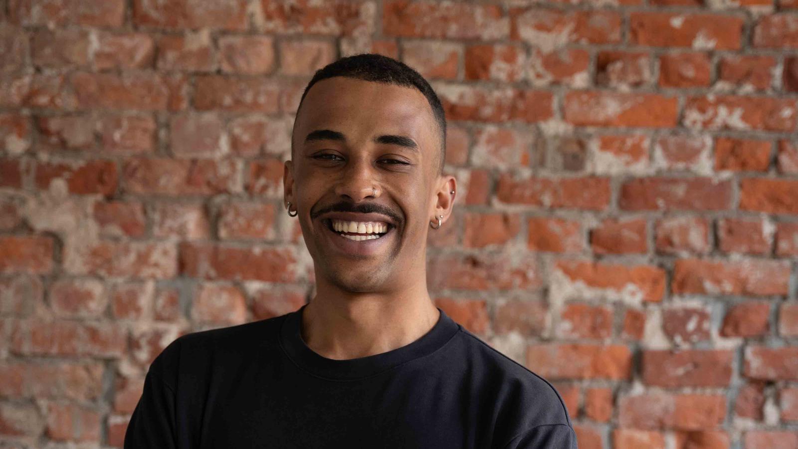 Smiling person with short hair and tattoos, wearing a black shirt, stands against a textured brick wall.