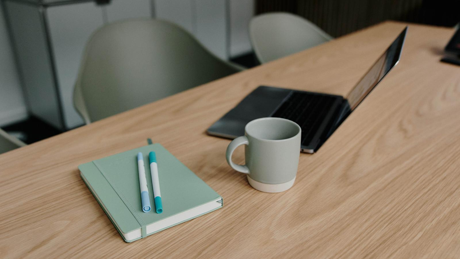 Notebook with pens, a mug, and an open laptop on a wooden desk, creating a minimalist workspace.